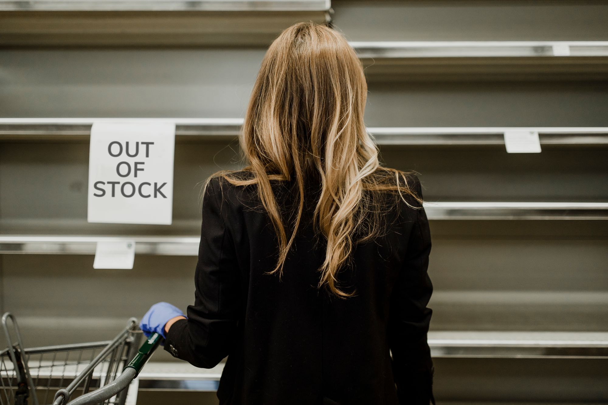 A blonde-haired woman stands in front of empty shelves, her expression reflecting concern and uncertainty.